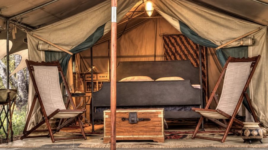 The inside of a canvas safari tent, with a bed, a writing desk and a lantern lit