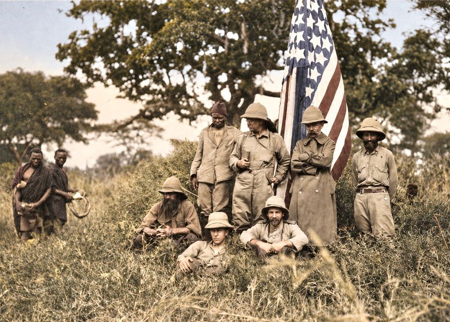 An early twentieth century safari party posed in tall grass beside a United States flag